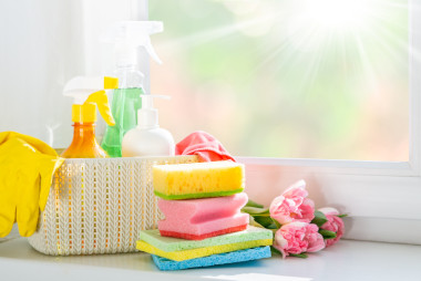 rayon de soleil qui éclaire à travers une fenêtre des fleurs roses à côté d'un panier blanc contenant des produits de nettoyage et des éponges de différentes couleurs